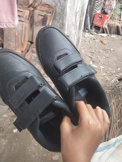 In Kedung Dowo, Indonesia, a close-up shot focuses on a pair of black, velcro-strap children's shoes being held by a person's hand. The shoes appear to be school shoes, with a matte finish and visible stitching. The inner sole of one shoe is partially visible, revealing a label with text that reads "HOMY KID SMART" and "150.000". The background consists of a pile of reddish-brown roof tiles and a rough, textured wall. Further in the background, there is a red plastic crate and dry vegetation. The ground is made of dirt and debris. The lighting suggests it is daytime.