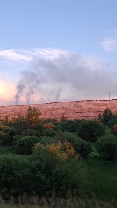 This media file depicts a landscape scene, likely taken from a moving vehicle due to the motion blur. The image is oriented vertically, with the sky occupying the left half and the ground the right. The sky is a pale blue with scattered, wispy white clouds that are tinged with pink and orange hues, suggesting it is either sunrise or sunset.
The right side of the image shows a densely wooded area with various shades of green foliage, interspersed with a few trees displaying autumn colors of yellow and orange. The ground appears to be a mix of grass and possibly some undergrowth. In the middle ground, a long, flat structure covered in a textured, reddish-pink material is visible. This structure runs horizontally across the frame and appears to be a man-made object. A single lamppost stands at the far end of this structure.
The overall mood is serene, with the soft colors of the sky and the natural elements of the trees. There is no visible text in the image. The setting is likely rural or semi-rural, given the open space and presence of trees. The time of day is either dawn or dusk.