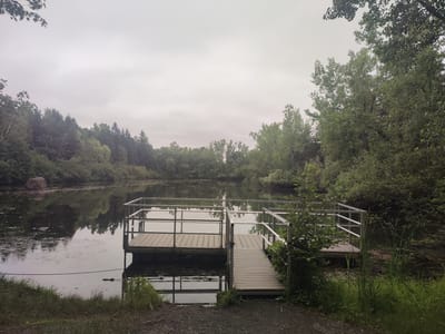 The image depicts a serene lake with a wooden dock on the right side, situated in the Parc national du Mont-Saint-Bruno in Saint-Bruno-de-Montarville, Canada. The sky is overcast, casting a muted light on the scene. Dense trees line the opposite shore of the lake, their reflections visible on the water's surface. A large, dark boulder is prominent on the left bank, also reflected in the water. The dock, made of light-colored wood planks, features metal railings and a ramp leading to the shore. Lush green vegetation, including tall grasses and bushes, surrounds the dock and the edge of the lake. The ground in the foreground is covered with gravel. The overall impression is one of a peaceful, natural setting, likely a popular spot for outdoor recreation.