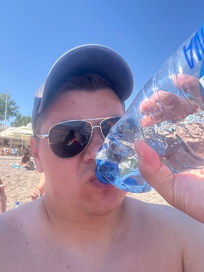 A man, wearing sunglasses and a cap, drinks water from a clear plastic bottle. He is shirtless and on a beach, as evidenced by the sand and people in the background. The scene appears to be outdoors under a clear blue sky. The background shows a beach with umbrellas and people lounging. The location is the city of Prijevor, Montenegro.