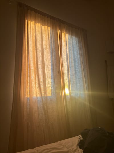 A low-angle, indoor shot shows sunlight streaming through a set of closed horizontal blinds. The blinds are a light tan color, and the sunlight casts a warm, yellow glow across them. Through the gaps in the blinds, hints of a blue sky are visible. To the right of the blinds, part of a bed is visible with a white comforter. A dark-colored garment hangs over the side of the bed. The walls are a light neutral color, and the ceiling appears to be white. The image does not contain any elements that indicate its location is Maillane, France. There are no people, subjects, actions, or interactions captured in the scene.