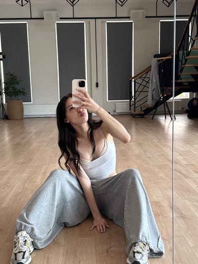 A young woman with long, dark hair is sitting on a wooden floor in what appears to be a dance studio or a gym. She is taking a selfie with her phone, which she is holding up to her face. She is wearing a white tank top and loose-fitting gray sweatpants. Her legs are spread wide apart, and she is looking at the phone with her lips puckered. In the background, there are large windows with blinds, a plant in a basket, and a staircase. The lighting is bright, and the overall scene suggests a casual and relaxed atmosphere. The location is Agalarov Estate, Russia.
