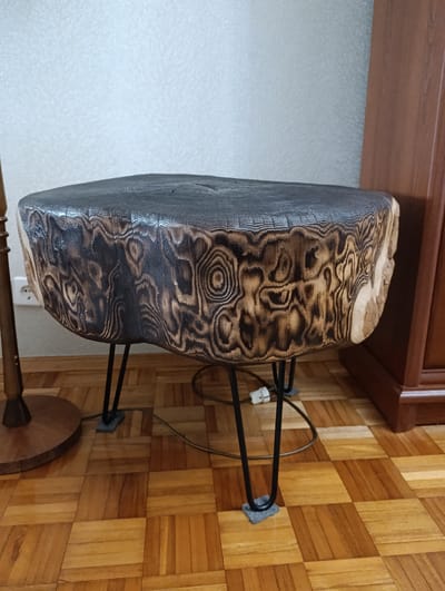 The image shows a handcrafted wooden stool sitting on a parquet floor. The stool's top is a dark, irregularly shaped slab of wood with a charred surface. The sides display a contrasting pattern of light and dark wood grain. Four black, hairpin-style metal legs support the stool, each with a small, gray pad at the bottom. The stool is positioned near a dark wood cabinet and a lamp with a rounded base. An electrical cord and plug are visible on the floor near the stool's legs. The background consists of a light-colored wall. The location is a room, presumably indoors. There is no indication this is in the city of Barvinok, Ukraine.