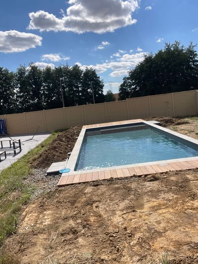 The image depicts an outdoor scene in Viry-Noureuil, France. The central focus is a swimming pool with clear blue water, bordered by a light-colored coping and a wooden deck. In the foreground, the ground is uncultivated dirt with tire tracks, suggesting ongoing construction or landscaping. To the left of the pool, a paved area is visible, with part of a metal lounge chair and a blue screen or cover. Beyond the pool and paved area, a light-colored fence stretches across the background, with a line of dense green trees behind it. The sky above is blue with scattered white clouds.