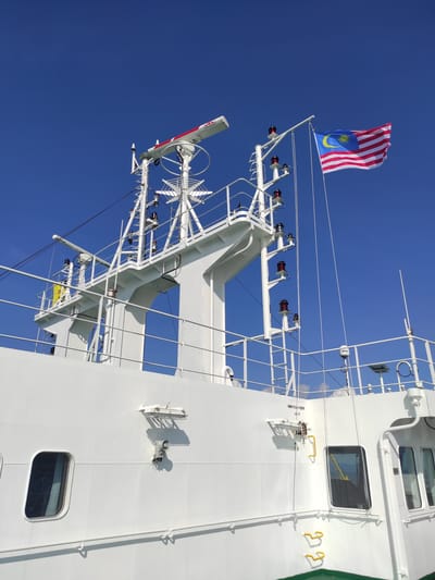 The image captures a partial view of a white ship's mast and superstructure against a clear, deep blue sky, suggesting bright daylight and fair weather. The scene is oriented from a low-angle perspective, looking upwards at the vessel.

Dominating the upper left of the ship's mast is a Malaysian flag (Jalur Gemilang), unfurled by the wind, showcasing its red and white stripes, a yellow crescent moon, and a 14-point star on a blue canton. Below the flag, the mast is adorned with various maritime equipment, including multiple antennas, a white radar scanner with red markings, and several red navigation lights mounted on white support structures and railings.

The ship's superstructure is painted a clean white, featuring rectangular porthole-style windows with dark, reflective glass. Various pipes, cables, and handholds are visible along its side, including a set of yellow, U-shaped handles. Shadows cast by the ship's components are sharp and distinct, indicative of direct sunlight.

Visible text details include "NAV. EQUIP. PSTR. VENT" stenciled near a vent on the ship's superstructure, and the word "SAILOR" on a white cylindrical antenna. The overall impression is of an active and well-maintained vessel operating under sunny skies, in the region of Kunak, Malaysia.