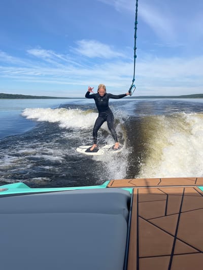 A young female is wakesurfing on a calm body of water. She is wearing a black wetsuit and is balancing on a white wakesurf board. She is holding onto a blue rope with a handle. The water is disturbed by the wake of a boat, creating white foam and splashes. The background shows a treeline and a blue sky with wispy clouds. The foreground shows part of a boat with grey seating and a brown deck. The image appears to have been taken on a sunny day. The location is Gromovo, Russia.