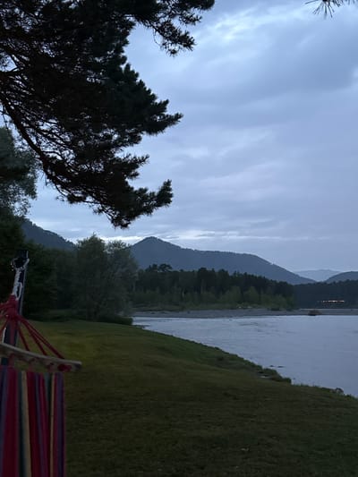 The image depicts a serene natural landscape, likely in Ust-Sema, Russia, during the twilight hours. The scene is dominated by a body of water, possibly a river, reflecting the overcast sky and the silhouettes of distant mountains. The mountains themselves are layered, with the closest ones covered in dense forest, while the furthest are shrouded in a soft haze.
In the foreground, a large, dark-leaved conifer tree stands prominently on the left, its branches reaching towards the sky. To the right, a gently sloping, lush green grassy hill leads down towards the water. At the very bottom of the frame, a colorful hammock, featuring stripes of red, yellow, blue, and purple, is visible, suggesting a place for relaxation.
The time of day is indicated by the soft, diffused light and the muted colors, suggesting either early morning or late evening. The weather appears to be cloudy, with streaks of lighter clouds against a pale blue sky. The overall mood is peaceful and tranquil, inviting a sense of calm. There is no visible text in the image. The image is oriented vertically.