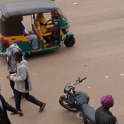 An elevated, eye-level, medium shot captures a street scene with several individuals and a three-wheeled auto-rickshaw. In the foreground, a person wearing a dark jacket and a purple beanie is partially visible, facing to the right. To the left, a motorcycle with a black seat and saddlebag is parked, facing forward, with its handlebars and instrument cluster visible.
Behind the motorcycle, two young men are walking. The one closer to the camera wears a grey hooded sweatshirt with black accents and black pants, and is facing left, with his hands held out. The other young man, to his left, wears a similar grey hooded sweatshirt with a dark design on the front and a red beanie, and is looking down.
The auto-rickshaw is a prominent feature in the background, with a dark grey canopy and a green and yellow body. A driver wearing a yellow long-sleeved shirt with black stripes is seated in the front, operating the vehicle. A passenger in blue pants is seated next to the driver. The auto-rickshaw has the number "19" painted on its front. The ground appears to be a dusty, reddish-brown paved surface, with scattered small pieces of paper. There is no indication of the location being Kayseri, Turkey.