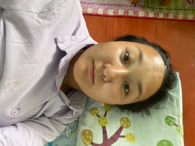 A close-up, eye-level, slightly high-angle, over-the-shoulder shot shows a person lying on their side on a light blue pillow with a cartoon tree pattern. The person is facing the camera and appears to be a young female with dark hair and brown eyes. She is wearing a light purple, collared shirt with buttons. Her head rests on the pillow. The background consists of a reddish-brown floor and a patterned rug. The scene is indoors, likely in a residential setting. There are no clear indicators of the specific city within Kyrgyzstan.