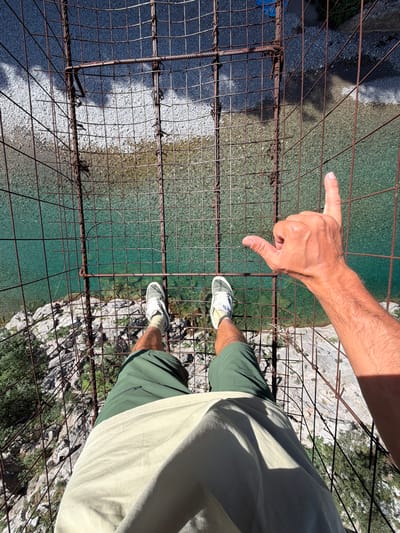 The image presents a first-person, downward-facing perspective of an individual standing on an aged, rusty metal grid structure. The subject's lower torso is visible, clad in light-colored shorts or fabric, with their legs wearing dark green shorts and white athletic shoes featuring light-colored soles and laces. The subject's right arm is extended, and their right hand forms a "shaka" gesture, with the thumb and pinky finger extended and the three middle fingers curled into the palm.

The metal grid beneath the subject's feet is composed of intersecting, heavily rusted rebar and thinner metal wires, forming predominantly square apertures. Below this grid, a body of exceptionally clear, turquoise-green water is prominently visible, revealing a bed of light-colored gravel and pebbles. To the left and directly beneath the subject's stance, a steep, rugged rocky bank or cliff face descends towards the water. This terrain is characterized by grey and white stones, interspersed with sparse green vegetation, consistent with scrub or small bushes.

The scene is illuminated by strong natural sunlight, casting distinct shadows. The metal grid casts a complex pattern of shadows onto the water and rocky ground below. The subject's right hand also casts a shadow onto their upper thigh/lower torso.

While the geographical features, including clear water, rocky terrain, and a suspended structure, are consistent with natural landscapes found in Montenegro, such as canyons or river valleys, the specific city within Montenegro cannot be identified from the visual information provided.