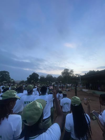 In the city of Emure-Ekiti, Nigeria, a large group of young adults, likely participants in the National Youth Service Corps (NYSC) program, are gathered outdoors at dusk. They are predominantly dressed in white t-shirts with green trim and green caps. Many are holding up their mobile phones, some filming or taking pictures of an unseen event or spectacle. The sky is overcast with a twilight hue, and trees and a covered structure are visible in the background, suggesting an open field or gathering area. The mood appears to be one of anticipation or observation, as the individuals are looking towards a common point of interest.
