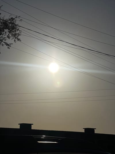 This is a vertically oriented photograph taken in Yablunivka, Ukraine, depicting a hazy sky with the sun prominently featured. The sun is a bright, almost blinding orb, emitting a strong lens flare that creates a vertical streak of light extending upwards. A smaller, more diffuse lens flare is visible to the right of the sun. A silhouette of a tree with sparse foliage is visible in the lower left corner. A complex network of power lines crisscrosses the sky, forming a stark, dark pattern against the bright background. On the right side of the image, a dark silhouette of a building facade is visible, featuring two rectangular protrusions, possibly vents or architectural elements, and a faint reflection of light on its surface. The overall atmosphere is muted due to the haze, suggesting either a foggy or overcast day, or possibly the effect of dust or pollution in the air. The time of day appears to be either early morning or late afternoon, given the angle and intensity of the sunlight. No people are visible in the image.