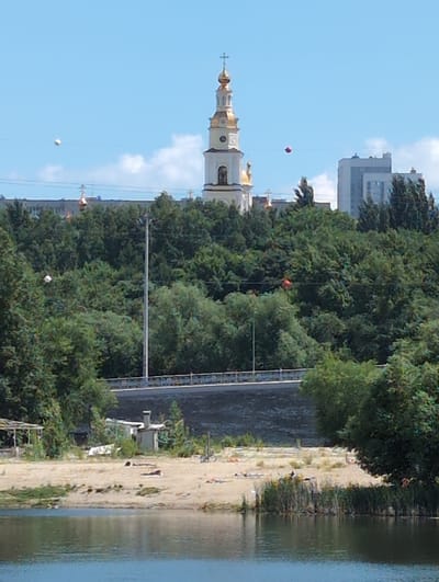 The image shows a view of the city of Vyshka, Russia, with a prominent church steeple and several buildings in the background. The foreground features a sandy riverbank with some vegetation and what appears to be a small structure. The river itself occupies the bottom portion of the image, reflecting the sky. In the middle ground, a dense line of trees separates the riverbank from the urban landscape. Overhead, two balloons are visible, one white and one red, suspended by strings. The sky is blue with scattered white clouds. The overall impression is a sunny day with a mix of natural and urban elements.
