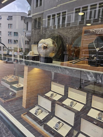 This image shows a street-level view of a luxury watch and jewelry store's display window, with reflections of the street and buildings visible. The primary subjects are the watches displayed in individual boxes, most of which are branded "PATEK PHILIPPE GENEVE" and one is branded "ROLEX". There are also necklaces and rings visible in the display. A large, metallic soccer ball is prominently featured, reflecting the surrounding environment. The scene is likely set in a city, given the architecture of the reflected buildings, and the presence of a store suggests a commercial area. The lighting suggests it could be daytime. There is text visible on the watch boxes and on a sign above the display, reading "ROLEX" and "SPRE". The overall impression is one of affluence and high-end retail.