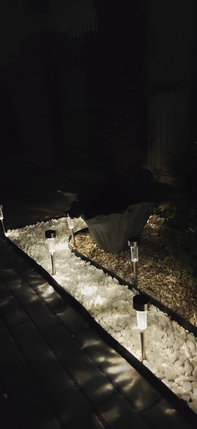 This is a nighttime image taken in the city of Pleszew, Poland. The scene depicts an outdoor pathway and garden area illuminated by solar-powered stake lights. The pathway appears to be paved with dark rectangular stones or tiles, and it runs along the edge of a garden bed filled with white pebbles. Several solar lights are placed within the white pebble bed, emitting a bright white light. To the right of the white pebble bed, there is another area of tan-colored pebbles and some dark foliage. In the background, there is a dark area with some vertical elements that suggest a fence or structure, and further in the background, a larger dark shape is visible, possibly an object or a shadow. A piece of light-colored fabric or material is draped over something in the mid-ground.