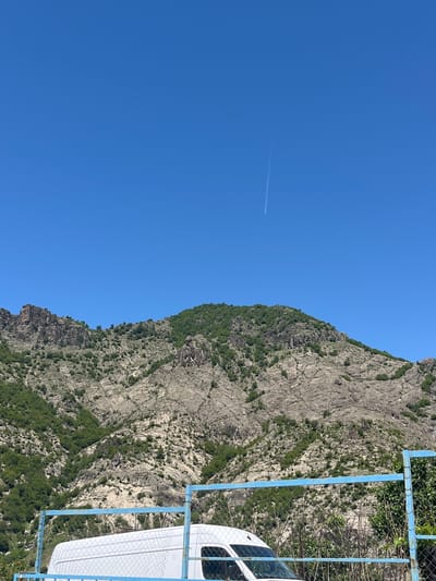 The image presents a bright, clear blue sky dominating the left half, with a contrail visible. A rugged, rocky mountain occupies the right side of the frame, sparsely vegetated with green trees. In the foreground, a section of a blue metal fence is visible, along with a portion of a white van, suggesting a roadside location. There is no definitive information to confirm its location within Bukovo, Bulgaria. The scene appears to be taken during a sunny day with a clear atmosphere.