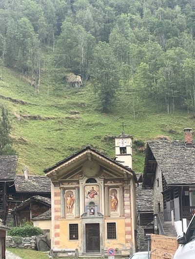 The image shows a quaint village nestled in the mountains of Alagna Valsesia, Italy. In the foreground, a small church with a yellow facade stands prominently. The church features religious frescoes on its exterior, and a bell tower with a clock rises beside it. The architecture of the surrounding buildings, with their slate roofs and stone walls, is typical of the Walser style. Behind the village, a lush green mountainside is covered with dense forest. The terrain is steep, with terraced fields and some evidence of past landslides or debris piles. The overall scene evokes a sense of peaceful seclusion and natural beauty.