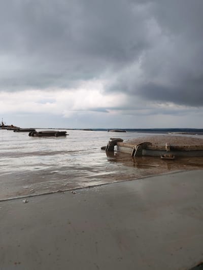 The image shows a flat, wet surface, likely a deck, covered with shallow water and possibly recent rain. Several rectangular, metal hatches with rusted hinges are visible, some partially submerged in the water. In the background, a dark, cloudy sky looms over a distant horizon, suggesting overcast or stormy weather. The scene appears to be outdoors, with a sense of industrial or maritime infrastructure, though no specific landmarks of Shchetinskoe, Russia are identifiable.