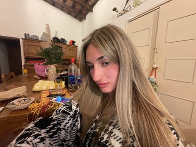A young woman with long blonde hair is taking a selfie in a room. She is wearing a black and white patterned jacket. On a table in front of her, there is a smartphone, an ashtray with cigarette butts, a yellow checkered cloth, a plant in a pink pot, and several bottles of water. In the background, there is a wooden cabinet with various items on top, including helmets and decorative objects. The room has a wooden ceiling and beige doors. The scene appears to be indoors, possibly in a residence. The image does not provide any information about the city of El Rodeo, Argentina.