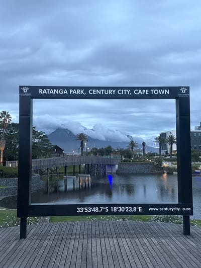 The image captures a scenic view framed by a black, rectangular sign. The sign is inscribed with the words "RATANGA PARK, CENTURY CITY, CAPE TOWN" and includes the Century City logo. On the opposite side of the sign, there is geographical information that says "33°53'48.7″S 18°30′23.8"E www.centurycity. o.za".
Through the frame, a landscape is visible, featuring a body of water with a wooden walkway extending across it. Palm trees are interspersed along the water's edge. In the background, a mountain range is partially obscured by clouds. The sky above is overcast. The scene appears to be taken in Ratanga Park, Century City, located in Milnerton, South Africa.
The foreground shows a wooden surface, adding depth to the image.