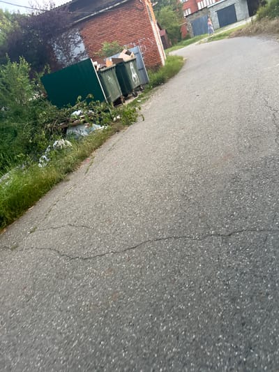 The image shows a rural road in the city of Apsheronsk, Russia, with a person walking in the distance. On the left side of the road, there are two large green trash receptacles and a dark green fence enclosing a small area. Scattered around the trash receptacles and in front of the fence is a pile of garbage, including cardboard boxes, plastic bags, and branches. Further to the left, there is dense green foliage. The road itself is paved with asphalt and shows signs of wear, with visible cracks. In the background, there are several buildings, including a brick structure and a gray garage with a large door. The sky is overcast.