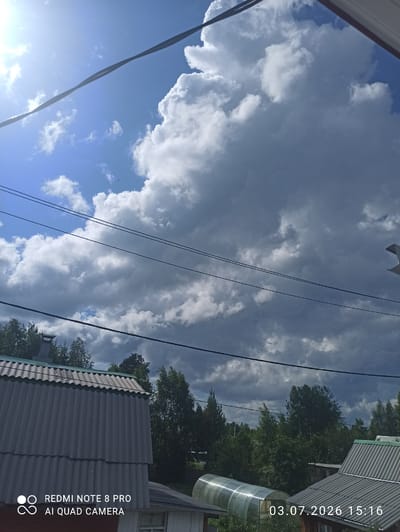 The image depicts an outdoor scene with a vast, cloudy sky. The clouds are a mix of dark and light gray, with patches of bright blue sky visible. The sun is shining brightly from the upper left corner, casting a lens flare across the sky. Several power lines are stretched across the frame, with one prominent line crossing diagonally from the top left to the bottom right. Below the sky, a line of trees is visible, with dense green foliage. In the foreground, the corrugated gray roof of a building is prominent, with a section of a greenhouse visible to the right. The image includes a timestamp in the lower right corner: "03.07.2026 15:16" and a watermark in the lower left corner indicating "REDMI NOTE 8 PRO AI QUAD CAMERA". The scene appears to be a residential area, possibly in a rural or suburban setting, given the presence of trees and simple structures. The city of Sovetskiy, Russia is not directly identifiable from the visual elements in the image.