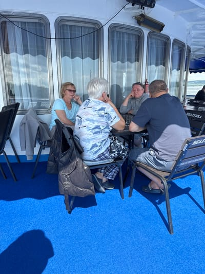 Four people are seated around a table on a deck of a ship. Two women and two men are present. The woman on the left is wearing a blue shirt and glasses. The woman next to her is wearing a patterned shirt. The man on the right is wearing a dark blue t-shirt and shorts, and the man next to him is wearing a grey sweater. The table has a dark surface and there are drinks on it. The deck is covered in bright blue carpet. There are large windows with white curtains behind the people. The ship appears to be on a body of water. The environment suggests a leisure activity on a sunny day. The specific location in Orel, Russia cannot be determined from the image alone.