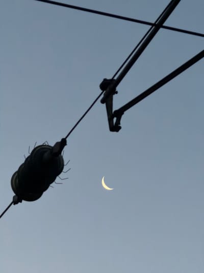 The image depicts a crescent moon in a pale blue sky, positioned to the right of a dark, overhead electrical wire infrastructure, likely related to a railway or tram line. To the left, a dark, cylindrical object with radiating elements, possibly an insulator or a part of the electrical system, hangs from a wire. The overall impression is one of urban infrastructure against a twilight or early evening sky, with a celestial element present. This scene is typical of the environment found near railway lines in Witten, Germany, where such overhead electrical systems are common.