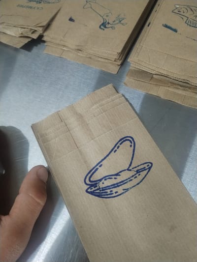 The image shows a stack of brown paper bags with blue prints of various sea creatures. In the upper left is a stack with a squid print. In the upper right is a stack with a fish print. The main focus is a stack of bags in the center with a blue print of an open clam. A finger rests on the edge of the table near the stack. The background is a gray metal surface. The bags possibly contain fish or other seafood based on the prints. The location is suggested as Pionersky, Russia, but there is no specific visual information in the image to confirm the location.