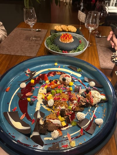 A large, blue plate showcases a dessert arrangement featuring ice cream scoops, chocolate pieces, fruit slices, crushed nuts, and dollops of whipped cream, accented with red petals and streaks of sauce. A separate dish holds a dark, circular cake surrounded by green foliage and adorned with red berries. To the side, small crackers sit on a black plate. Two wine glasses and silverware are placed on brown placemats.