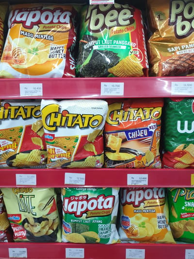 The image shows a display of potato chips on supermarket shelves. The shelves are red, and the products are arranged in rows. The lighting is bright, suggesting an indoor environment. The brands visible include Japota, Bee, Chitato, and Pocky. Several different flavors are advertised, such as Honey Butter, Grilled Seaweed, and Beef. Prices are displayed on white tags beneath each product. The overall scene is typical of a retail grocery store aisle.