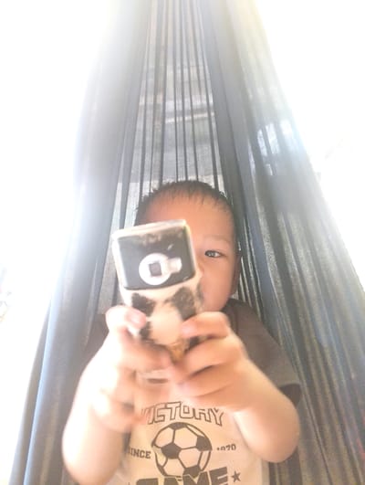 A young child is in a hammock, holding a cell phone up to the camera as if taking a selfie. The child's face is partially obscured by the phone and a dark mesh curtain. The child is wearing a light-colored t-shirt with a soccer ball graphic and the word "VICTORY" visible. The background consists of the hammock's dark mesh and bright light filtering through, suggesting an outdoor or well-lit indoor setting. The overall lighting is bright, with a halo effect around the child and phone. The image is slightly blurry, indicating motion or a quick capture. The location is described as Pompanua, Indonesia.