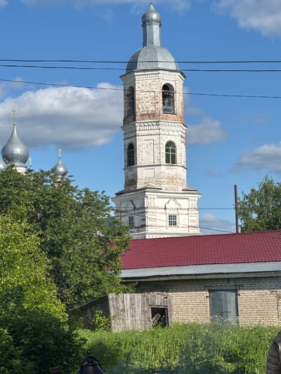 This media file depicts a serene, sunlit scene featuring a prominent religious structure in Луптюг, Russia. The main subject is a tall, weathered bell tower or church spire, constructed from white-painted brick with areas where the original red brick shows through. It has multiple levels, featuring both arched and rectangular windows, and is topped with a grey, metallic onion dome and a spire. Black power lines stretch across the upper part of the image, partially obscuring the tower and the sky.
To the right of the tower, a building with a distinct red corrugated metal roof is visible, suggesting an adjacent structure within the complex. Below it, there's a section of a light-colored brick wall with a metal covering, and an old wooden structure, possibly an outbuilding or shed, surrounded by overgrown green grass and wild plants. Lush green trees and bushes are scattered throughout the midground and foreground, partially obscuring other elements. In the lower left, behind some trees, several other grey, metallic church domes, each topped with a cross, are visible, indicating that the bell tower is part of a larger church or monastery complex.
The sky is a vibrant blue with scattered white, fluffy clouds, suggesting a clear and sunny daytime. There are no people visible, and no discernible text. The image's content is characteristic of traditional Russian rural or small-town architecture and natural landscapes. The entire image is presented as if rotated 90 degrees clockwise from its natural upright orientation.