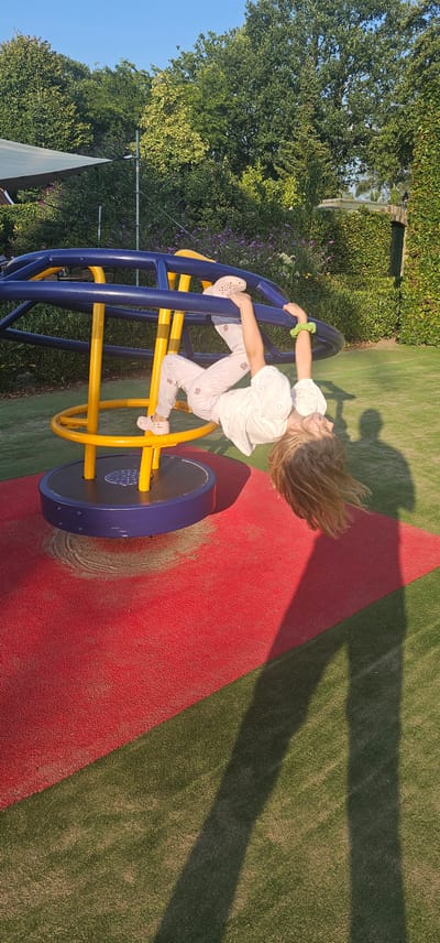A young child with blonde hair, wearing a white top and pink patterned pants, is upside down, hanging from a blue and yellow merry-go-round. The merry-go-round is on a red and green play surface in an outdoor setting. Green trees and bushes form a dense backdrop, with a clear blue sky above. The sun casts long shadows of the child and the merry-go-round onto the ground, suggesting it is late afternoon. The location is the city of Groenlo, Netherlands.