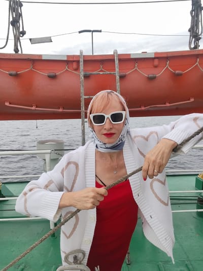 A woman with pink hair, wearing white sunglasses, a patterned headscarf, a white cardigan with heart designs over a red dress, and a necklace, stands on a boat deck. She is holding a thick rope with both hands. Behind her, an orange lifeboat is mounted. The background shows the ocean under a cloudy sky. The deck is green. The location is the city of Bereg, Russia.