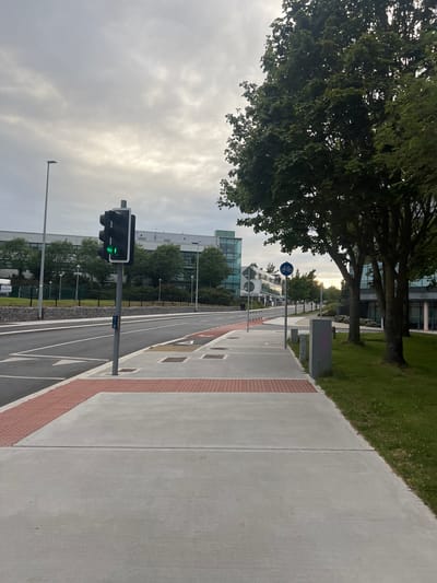 The image depicts a street scene in Balbriggan, Ireland, likely during overcast daylight. In the foreground, a concrete sidewalk runs alongside a paved road. The sidewalk has a textured red strip at its edge. To the right of the sidewalk is a grassy area with several mature trees.
On the sidewalk, there are several gray concrete bollards and a utility box. A traffic light is visible on a pole, displaying a green light. Next to the traffic light, a blue sign with a white bicycle symbol indicates a cycle path. The road is marked with white lines, and further down, red markings are visible, possibly indicating a cycle lane.
In the background, several modern buildings with large glass windows and trees are visible. The sky is overcast with diffused sunlight. The scene appears to be an urban or suburban environment with dedicated pedestrian and cycle infrastructure. There are no people clearly visible in the immediate foreground or midground of the image.