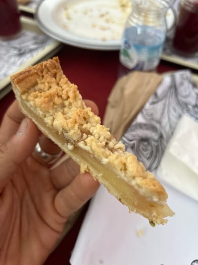 A hand holds a slice of crumble-topped pie, possibly an apple or pear crumble, in a close-up shot. The pie has a golden-brown crumble topping over a light-colored filling, with a flaky pastry base. A silver ring is visible on the ring finger. The background is blurred but suggests an outdoor or casual setting, with glimpses of a white plastic bottle, a patterned paper or cloth, and what appears to be a dark red surface, possibly a tablecloth. The lighting suggests daytime. There is no visible text in the image.