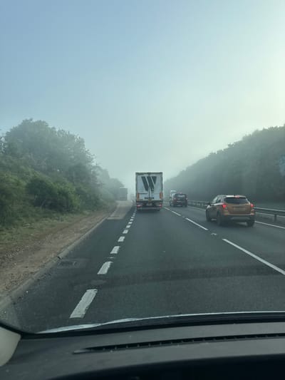 The image shows a foggy highway scene in Babergh, United Kingdom. Several vehicles, including a large truck and a gold SUV, are visible on the multi-lane road. The environment is obscured by heavy fog, reducing visibility. Trees line the side of the road. The photo appears to be taken from inside a vehicle.