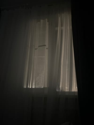 An interior scene captured in low light features a window covered by sheer, white or light-colored curtains. The curtains are drawn across the entire window, exhibiting vertical folds. A darker, opaque curtain or drape is visible partially open on the far right side of the frame, creating a vertical dark block. A diffused light source from outside the window illuminates the sheer curtains, casting a soft glow into the otherwise dim room. Faint details or textures, possibly condensation or dirt, are discernible on the windowpane through the translucent fabric. The room's interior on the left and bottom is dark, devoid of visible objects or individuals. The location is Dmitriyevskaya, Russia.