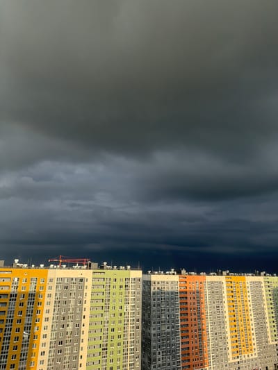 The image shows an exterior view of a tall, modern apartment building in the city of Анкудиновка, Russia, contrasting with a dark, cloudy sky. The building's facade is characterized by horizontal bands of different colors – yellow, gray, orange, green, and white – giving it a vibrant, contemporary look. Numerous windows are visible on each floor, suggesting a high density of residential units. A construction crane is attached to the side of the building, indicating ongoing development. The sky is dominated by dark, ominous clouds, suggesting an impending storm.