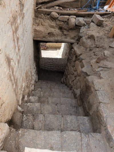 A stone staircase descends into a shadowed opening in a rough-hewn stone and concrete structure. The walls are weathered and uneven, with some sections appearing plastered and stained. Debris, including rocks, wood, and what appears to be blue netting, is visible in the background above the opening. The lighting suggests an outdoor or semi-enclosed environment. This scene is located in Choma, India.