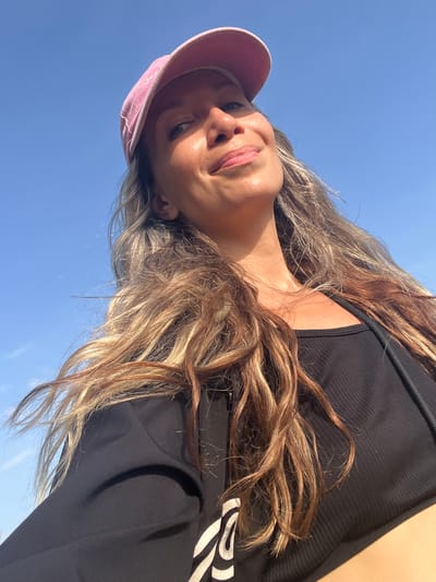 A low-angle, close-up selfie is taken by a woman wearing a pink baseball cap and a black crop top. The woman's face is turned upwards and to the right, with her eyes looking towards the camera. Her hair, which is long and wavy with highlights, is blowing in the wind. The background is a clear blue sky. The image appears to have been taken outdoors, possibly in the city of Talalla, Sri Lanka, given the bright sunlight and clear sky.