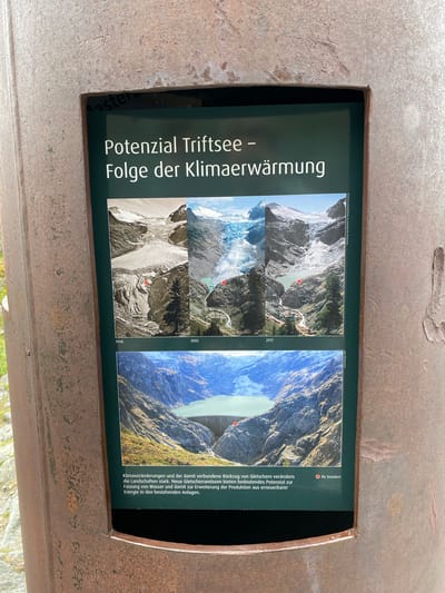 An outdoor informational display sign, framed by a rusted, weathered metal structure, is presented in a close-up, eye-level shot. The sign, positioned in Innertkirchen, Switzerland, features a dark green background with white German text and two main photographic sections.

The top section displays a triptych of three landscape photographs documenting glacier retreat. The leftmost image, labeled "1948," is a black-and-white aerial view of an extensive glacier system, with a red circular marker positioned on the lower part of the glacier. The central image, labeled "2002," is a color photograph from a similar vantage point, showing a significantly reduced glacier and a small, turquoise meltwater lake at its base, with a red circular marker in the vicinity of the lake. The rightmost image, labeled "2011," is also a color photograph, depicting further glacier retreat, a larger, more defined meltwater lake, and exposed rock formations, again with a red circular marker near the lake. Vertical text along the right edge of this section reads "Sammlung Gesellschaft für ökologische Forschung" (Collection Society for Ecological Research).

Below this triptych, a larger, single color photograph displays a wide view of a large, turquoise artificial lake nestled within a mountainous valley, with a substantial concrete dam visible across the valley. A red circular marker is placed on the surface of the lake, near the dam.

Beneath the lower photograph, white German text is inscribed: "Klimaveränderungen und der damit verbundene Rückzug von Gletschern verändern die Landschaften stark. Neue Gletscherrandseen bieten bedeutendes Potenzial zur Fassung von Wasser und damit zur Erweiterung der Produktion aus erneuerbarer Energie in den bestehenden Anlagen." (Climate changes and the associated retreat of glaciers are significantly altering landscapes. New glacier-fed lakes offer significant potential for water retention and thus for expanding the production of renewable energy in existing facilities.) To the right of this text, a small red dot accompanies the words "Ihr Standort" (Your Location), indicating the viewer's current position relative to the depicted scene.

Portions of green foliage are visible in the blurred background to the left of the metal frame, indicating an outdoor, natural setting.
