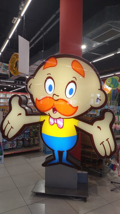 A large, illuminated, cartoon-like cutout of a man stands in the middle of a store. The man has a round, yellow head with orange eyebrows and ears. He has large blue eyes, a prominent orange mustache, and a pink and white striped bowtie. His body is yellow on top and blue on the bottom, resembling a shirt and pants. His arms are outstretched, and his hands are in a thumbs-up gesture. The cutout is mounted on a grey pedestal with wheels. In the background, shelves of products are visible, along with ceiling lights and a red pillar. The floor is tiled with a square pattern. The overall scene appears to be a retail environment, likely a toy store or a department store, within the city of Pylaia Municipal Unit, Greece, based on the provided information.