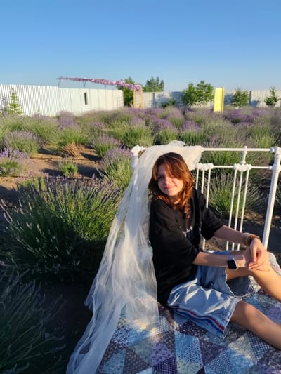 A young person with shoulder-length brown hair and light brown eyes is sitting on a white ornate bed frame in a lavender field. They are wearing a black t-shirt and light blue denim shorts with red and blue stripes on the side. A light white veil is draped over the bed frame behind and to the left of the person. The bed is covered with a colorful patchwork quilt. The person is looking at the camera with a slight smile, and their right arm is resting on their right leg, while their left arm is bent with their hand on their knee. A watch with a grey band is visible on their left wrist.
The scene is a lavender field under a clear blue sky. In the background, there is a white picket fence, some trees, and a decorative yellow door. The sun appears to be low in the sky, casting a warm glow on the scene, suggesting it is late afternoon or early evening. The overall mood is peaceful and idyllic.
Nechaiane, Ukraine is the stated location context.