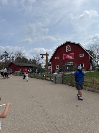 A public outdoor space near "The Barn" in Powell, Ohio, United States, shows a red barn building with white trim under a cloudy sky. A person in a blue shirt and white cap is seen walking along a paved path, next to a wooden fence. People are scattered throughout the area, some near another red building further down the path. Trash and recycling bins are present.