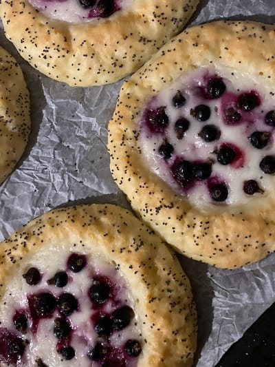 This is a close-up, overhead shot of three berry and cream pastries. The pastries have a golden-brown, slightly puffed crust sprinkled with poppy seeds. Each pastry has a central depression filled with a creamy white filling and whole dark berries, likely blackcurrants or blueberries, with some juice staining the cream a purplish hue. The pastries are arranged on crumpled grey parchment paper, which is on a dark surface. The lighting is somewhat dim, creating shadows and highlighting the textures of the food and paper. This image does not contain any identifiable landmarks or elements that would allow for its specific location in Velykyi Bychkiv, Ukraine to be determined.