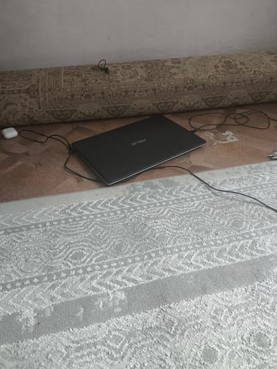The image shows a laptop, an earbud charging case, and several cords on the floor, partially covered by a rolled-up rug and a large, light-colored patterned rug. The laptop is closed and appears to be black with "ASUS" visible on its surface. The earbud case is white. The background consists of a rolled-up rug with a traditional pattern, and the floor is a textured brown material. The foreground is dominated by a large, light-colored rug with an intricate, repeating pattern. The scene appears to be indoors, likely in a home or office setting. There are no people or clear indicators of the time of day or weather. The location context provided is Қаратөбе ауылдық округі, Kazakhstan, suggesting this is the geographical setting.