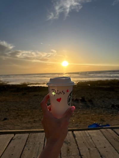 A Starbucks cup, labeled "Prins" with a heart and star, is held in someone's hand. The cup is positioned against a bright, setting sun reflecting on the ocean. To the right, a wooden boardwalk is visible. The background features a sandy or pebble-covered shoreline extending towards the horizon. The overall scene evokes a calm, beachside atmosphere.