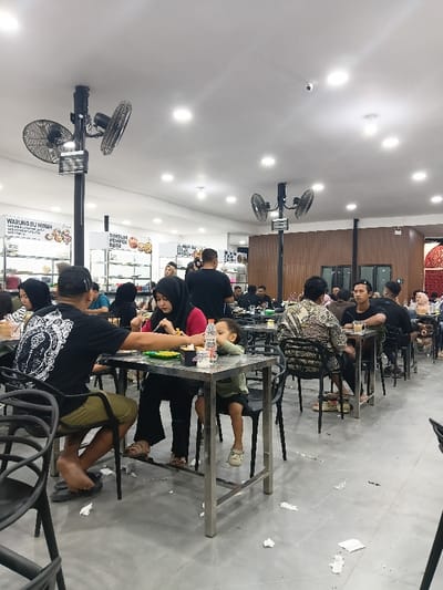 The image depicts an indoor, brightly lit food court or casual restaurant in Seumadam, Indonesia. Numerous patrons are seated at rectangular, metal-framed tables with black chairs, engaged in dining and socializing.

The environment features a white ceiling with multiple recessed LED lights and several black poles supporting dual-head industrial ceiling fans. The floor is light grey concrete or large tiles, with scattered white paper napkins. Signs in Indonesian advertising "WARUNG BU MINAH" and "DIMSUM PEMPEK DESE" are visible on the white walls behind glass display cases in the left background. A dark brown wooden panel wall occupies the right side of the dining area. A black security camera is mounted on the ceiling in the mid-ground.

In the foreground, a man in a black t-shirt with a white graphic print on the back and olive green shorts, wearing flip-flops, is seated with his back to the camera, reaching towards a plate. Opposite him, a woman in a red shirt and black hijab is actively eating from a plate with a fork. To her right, a young child in a light green t-shirt and dark shorts sits, looking towards the right, with a plastic water bottle on the table. Other individuals, predominantly adults in casual attire, some wearing hijabs, are scattered across the dining area at various tables, eating and drinking. A man in the mid-ground wears a distinctive patterned long-sleeved shirt.